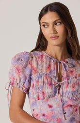 A woman is wearing a short-sleeved blouse with a watercolor floral pattern in shades of pink, purple, and peach. The blouse has gathered puff sleeves with ties at the cuffs, a ruffled yoke across the chest, and a front tie neckline.
